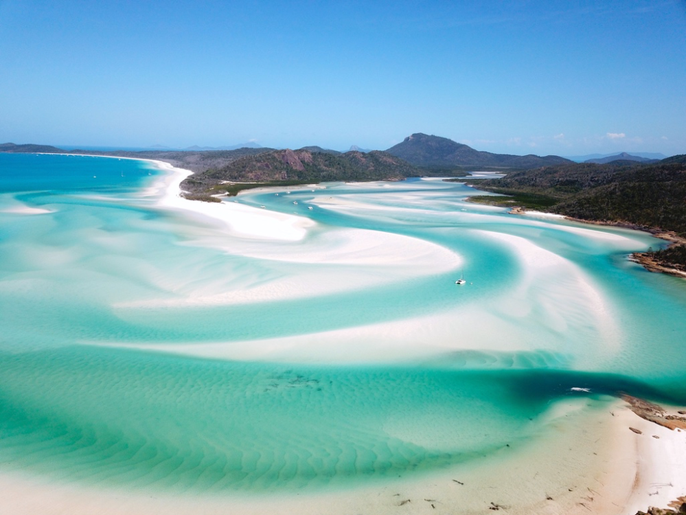 Aerial view of a turquoise beach with white sandbanks and distant hills under a clear blue sky.
