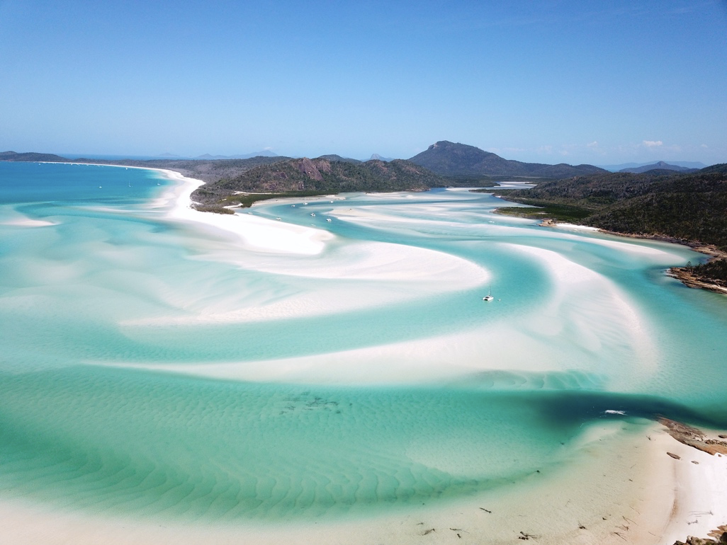 Aerial view of a turquoise beach with white sandbanks and distant hills under a clear blue sky.