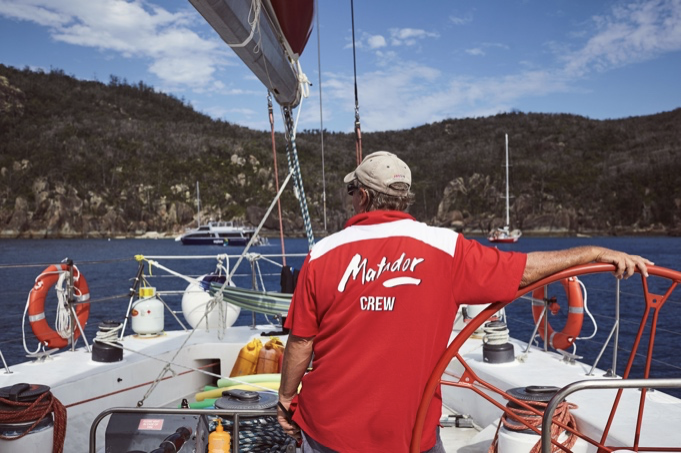 Person in red 'Matador Crew' shirt steering a sailboat with rocky coastline in the background.