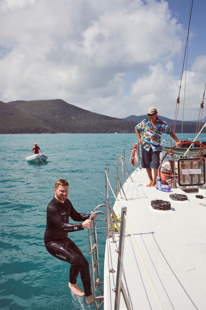 Man in wetsuit climbs ladder onto sailboat, another man stands on deck, with mountains and clouds in background.