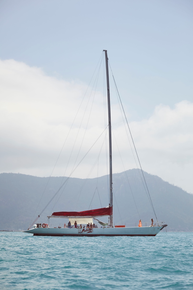 Sailboat with red sail and people onboard in blue sea with mountains in background.