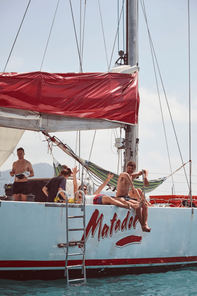 People relaxing on a sailboat with a red sail and the name 'Matador' painted on the side.