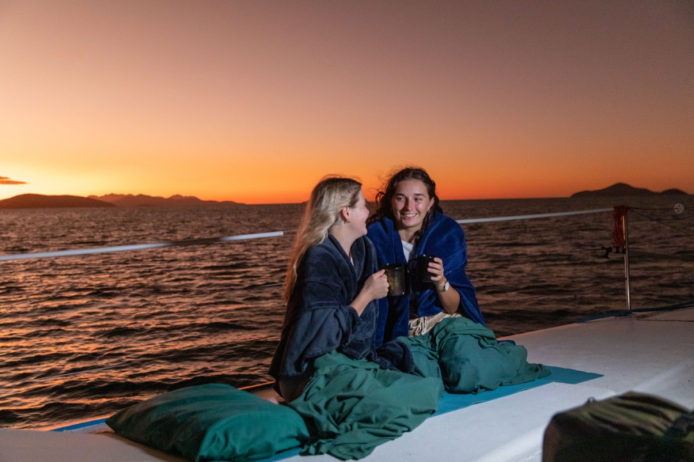 Two people wrapped in blankets on a boat at sunset, holding mugs and smiling.