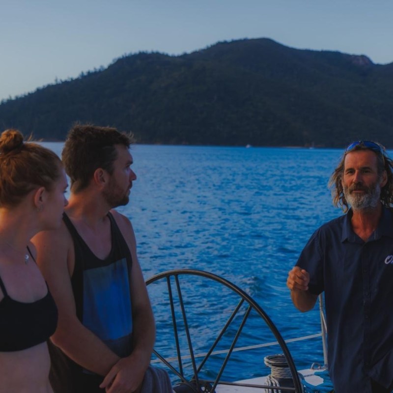 Three people on a boat, ocean and forested island in background, evening light.